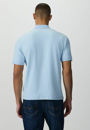 Muž v svetlomodrej textúrovanej polo košeli s krátkym rukávom a tmavomodrých džínsach, pohľad zozadu na neutrálnom pozadí.