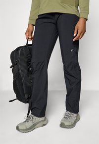 Black outdoor pants with a slim fit, made from lightweight material. Features a subtle logo accent and are paired with gray hiking shoes.