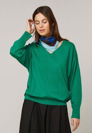 Femme en pull vert à col en V et jupe noire ajustant un foulard bleu à motifs sur un fond uni.