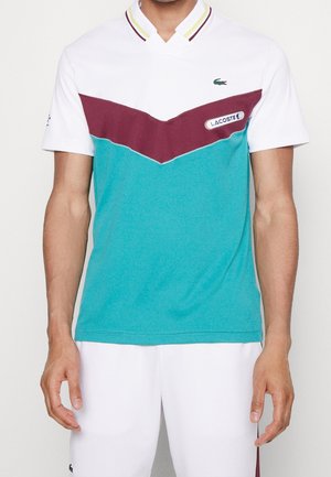 Mężczyzna noszący polo z krótkim rękawem w białe, bordowe i morskozielone paski w kształcie szewronu, z logo Lacoste na piersi, w połączeniu z białymi szortami.
