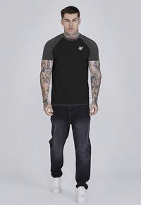 Schwarz-graues, kurzärmeliges T-Shirt mit Logo, kombiniert mit dunklen Jeans und weißen Sneakern. Das Model hat sichtbare Tätowierungen an den Armen.