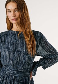 Mörkblå klänning med långa ärmar, med en texturerad tyg och ett mönster av vertikala linjer och blommiga detaljer. Åtsittande midja med en flödande kjol.