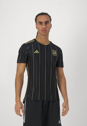 Jersey deportivo negro con rayas doradas verticales, presenta un cuello redondo, mangas cortas y un logo bordado en el pecho.