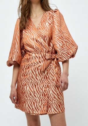 Kobieta w pomarańczowo-beżowej sukience z motywem zebry, z bufkami i trzy-czwartkowymi rękawami oraz kokardą zawiązaną w talii, stojąca na jednolitym tle.