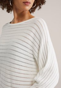 Pull blanc avec un motif à rayures horizontales et une texture en maille ajourée. Coupe ample avec manches larges et col rond.