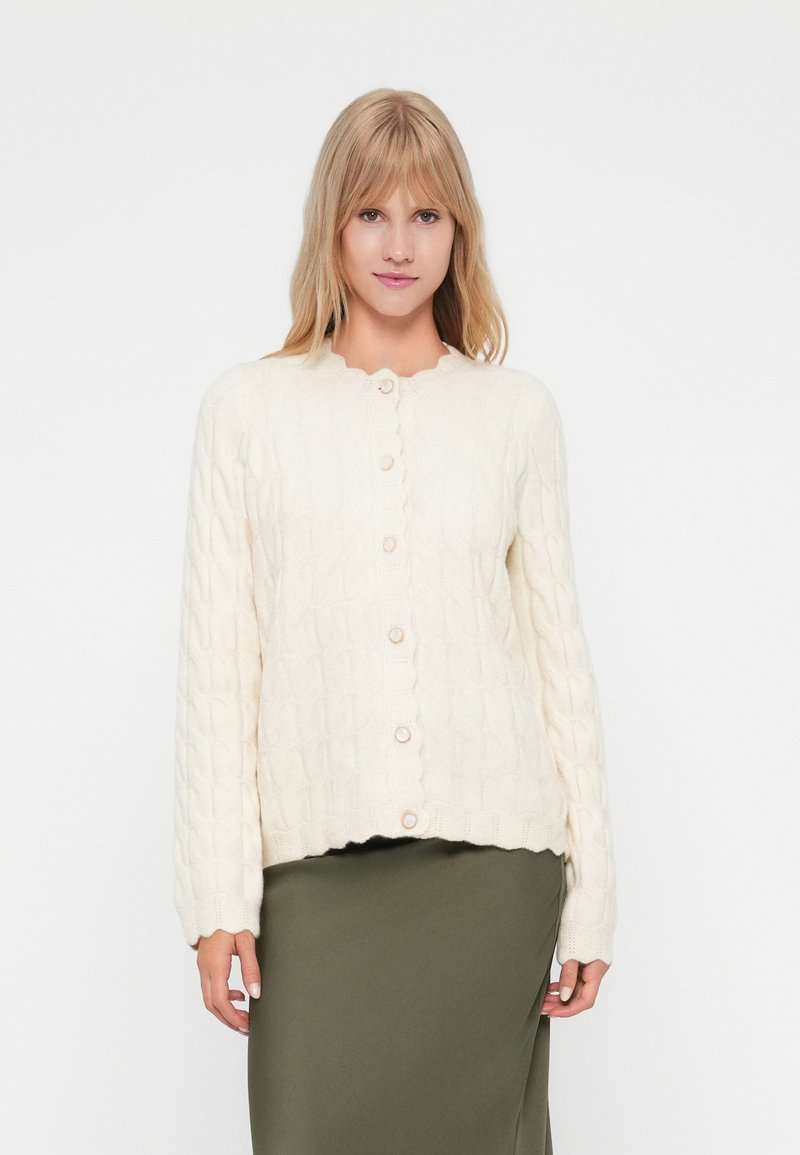 Kabelstrikket cardigan i creme, med en bølget krave, frontknaplukning og ribbede mansjetter, parret med en grøn nederdel.