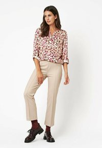 Blumenbluse mit pinken und burgunderfarbenen Mustern, kombiniert mit beigen maßgeschneiderten Hosen. Das Model trägt dunkle Schnürschuhe und burgunderfarbene Socken.