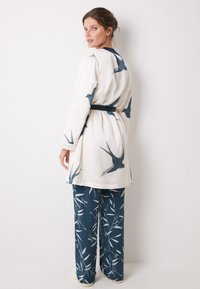 Robe bianca a maniche lunghe con motivo di uccelli blu, cintata in vita, abbinata a pantaloni navy a gamba larga con un design floreale.