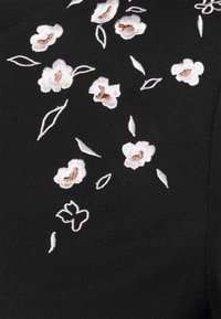 Svart tyg med vit och ljusrosa blommig broderi, med detaljerade blad och kronblad, vilket skapar en texturerad design.