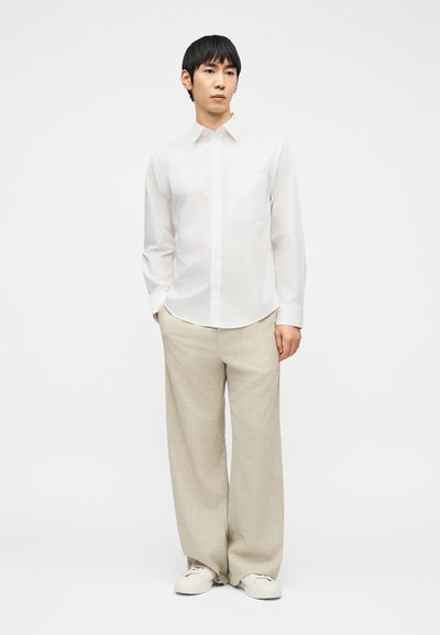 Νεαρός άνδρας που στέκεται φορώντας λευκό πουκάμισο με κουμπιά, μπεζ παντελόνι με φαρδιά βάση και λευκά αθλητικά παπούτσια, μπροστά σε μονόχρωμο φόντο.