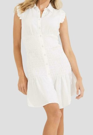 Femme portant une robe blanche sans manches boutonnée, taille smockée et ourlet à volants, se tenant devant un fond gris clair uni.