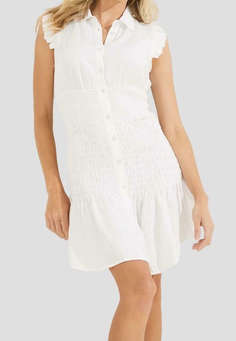 Femme portant une robe blanche sans manches boutonnée, taille smockée et ourlet à volants, se tenant devant un fond gris clair uni.