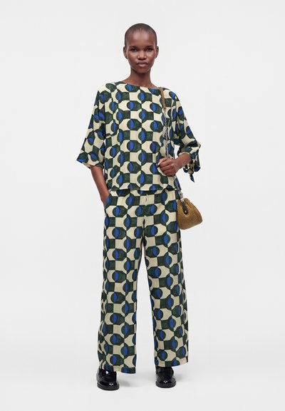 Femme debout portant un haut et un pantalon large assortis avec un motif géométrique vert, bleu et beige, tenant un sac à main tressé marron avec une bandoulière chaîne.