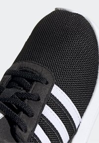 Svarta sneakers i meshmaterial, med vita ränder, texturerad ovandel och svarta snören. Sulan är vit och något upphöjd.