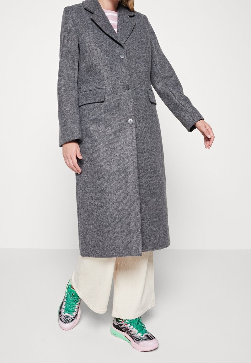Personne portant un long manteau gris en laine, un pantalon blanc large et des baskets colorées avec des lacets verts, debout devant un fond clair uni.