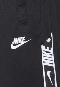 Czarne sportowe spodnie o gładkiej fakturze, z białym logo Nike oraz bocznym paskiem z powtarzającym się, wyraźnym nadrukiem marki.