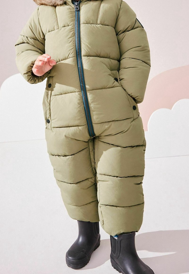 Enfant portant une combinaison d'hiver matelassée vert olive avec une fermeture éclair à l'avant et des bottes en caoutchouc noires, debout sur un sol clair.