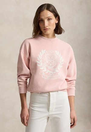 Žena s hnedými vlasmi po plecia nosí ružovú mikinu s bielym logom Polo Ralph Lauren Wimbledon a biele nohavice na neutrálnom pozadí.