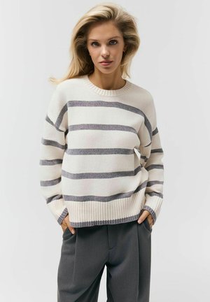 Blondynka z rękami w kieszeniach, ubrana w kremowy sweter w szare poziome paski i ciemnoszare spodnie na jednolitym tle.