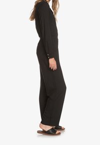 Mono negro con mangas largas, detalles de botones y bolsillos. Combinado con sandalias negras de piso plano y diseño de punta abierta.