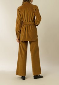 Ruskea corduroytakkia, jossa on vyöllinen vyötärö, pitkät hihat ja rento istuvuus, yhdistettynä samansävyisiin leveälahkeisiin housuihin ja mustiin kenkiin.