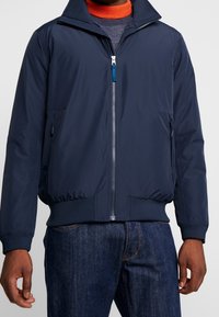 Navy blue lightweight jacket with a zip front, high collar, ribbed cuffs, and pockets. Smooth, water-resistant fabric with a fitted design.