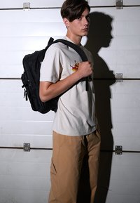 Mochila negra con múltiples compartimentos, llevada en el hombro derecho por una persona con una camisa beige clara y pantalones marrones, frente a una pared blanca.