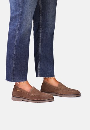 Personne portant un jean court bleu foncé et des mocassins en daim marron, debout sur un fond clair uni.