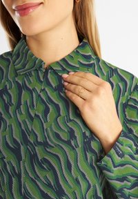 Camicia a maniche corte verde e blu navy con un motivo a onde e strisce bianche. Design del colletto con chiusura a bottone frontale. Anello dorato al dito.