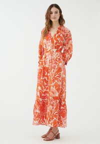 Abito maxi floreale in tonalità di arancione e rosa, caratterizzato da un design avvolgente, maniche lunghe, vita arricciata e orlo a strati. Abbinato a sandali marroni.