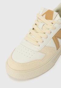 Kreemja ja beež sneaker, millel on sile nahk ja pehme seemisnahk, perforeritud varvas, ühtlased paelad ja geomeetrilised aktsendid küljel.