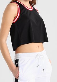 Musta lyhyt crop-top, jossa on verkkotekstuuri ja punavalkoiset raidat kaula-aukossa ja käsivarsien rei'issä. Yksinkertainen muotoilu, löysä istuvuus.