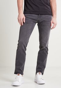 Jeans de mezclilla negra con un ajuste slim, con un lavado desgastado, diseño de cinco bolsillos y combinados con zapatillas blancas.