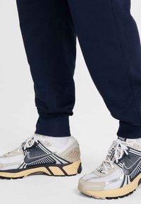 Tumši zili sporta bikses ar elastīgām aprocēm, kombinētas ar viegliem Nike sporta apaviem. Sporta apaviem ir tīkliņa augšdaļa, baltas, melnas un bēšas krāsas akcenti un teksturēta zolīte.