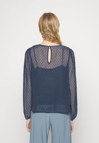 Blusa transparente azul-marinho com bolinhas texturizadas, mangas longas, fecho de botão nas costas em forma de fenda, e uma camada sólida suave por baixo.