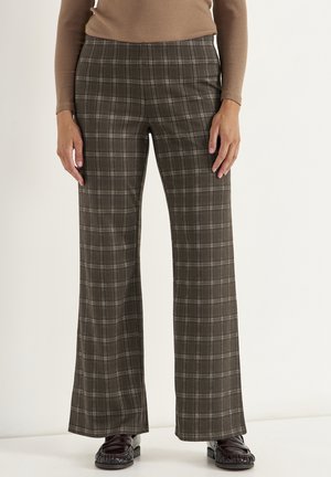 Person iført brune ternede bukser med vide ben, en langærmet beige top og sorte loafers, stående mod en ensartet lys baggrund.