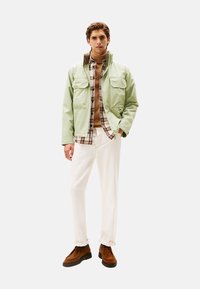 Chaqueta verde clara con dos bolsillos frontales, superpuesta sobre un suéter de cuello alto marrón y una camisa de cuadros. Combinada con pantalones blancos y zapatos marrones.