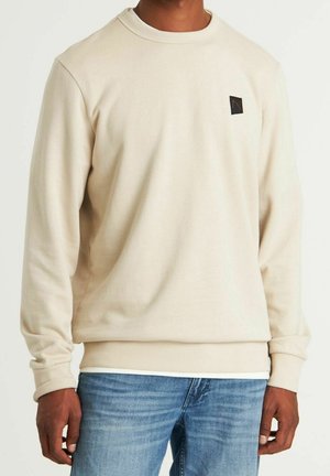 Mann trägt cremefarbenen Langarmpullover mit kleinem schwarzem Logo auf der Brust und hellblaue Jeans vor einfachem Hintergrund.