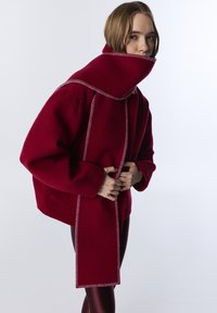 Červený kabát z vlněné směsi s vysokým límcem a velkým šátkem. Má kontrastní švy a boxy střih, spárovaný s lesklými tmavými legínami.