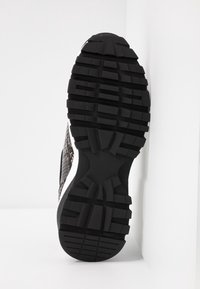 Semelle de chaussure en caoutchouc noir avec des crampons épais reposant contre un mur et un sol blancs, montrant un motif de grip détaillé.