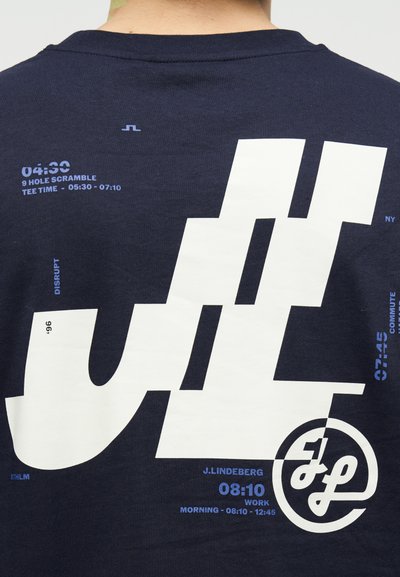 Μπλε πουκάμισο ναυτικού μπλε στο πίσω μέρος με μεγάλο λευκό στιλιζαρισμένο λογότυπο, μικρό μπλε κείμενο που απαριθμεί ώρες και λέξεις σχετικές με το γκολφ και τα ωράρια εργασίας.