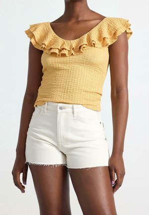 Femme portant un top jaune texturé à volants et épaules dénudées, associé à un short en jean blanc taille haute avec des ourlets effilochés.
