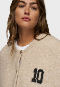 Joven mujer con cabello castaño claro lleva un cárdigan beige abotonado con el número negro "10" en el pecho sobre una camiseta blanca, fondo neutro.