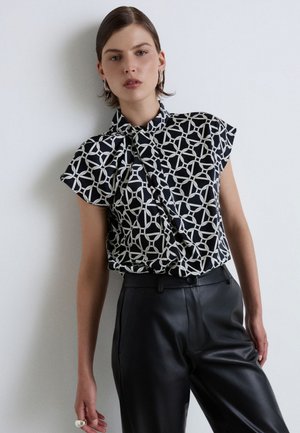 Donna con capelli corti che indossa una blusa senza maniche a motivo bianco e nero e pantaloni di pelle neri, in piedi contro un muro bianco.