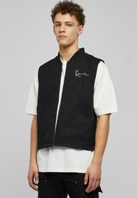 Melna bez piedurknēm veste no strukturēta auduma ar priekšējo rāvējslēdzēju, ar sānu kabatām un baltu firmas logo uz krūtīm.