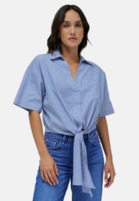 Camicia a maniche corte blu e bianca a righe con colletto, dettaglio a nodo in vita e vestibilità rilassata, abbinata a jeans in denim blu.