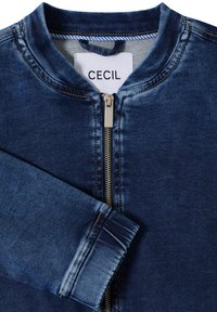 Veste en jean bleu foncé avec fermeture éclair et encolure ronde. Présente des détails de couture subtils et une texture lisse. Étiquette de marque visible.