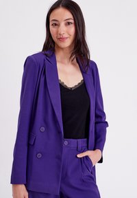 Blazer double boutonnage violet au design cintré, avec des poches avant et des boutons assortis, porté sur un débardeur en dentelle noire.