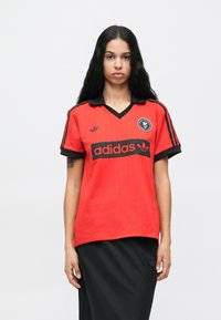 Czerwona koszulka Adidas z czarnymi akcentami, dekoltem w serek, krótkimi rękawami, z pionowymi paskami i logo na lewej piersi.