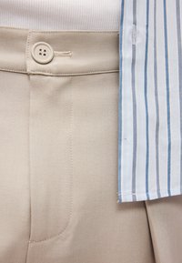 Beezhihallid, millel on sile pind ja nööbiga sulgemine. Kergelt sinise triibuga särk on kaelaosa nähtav, tuues sisse kontrastset mustrit.